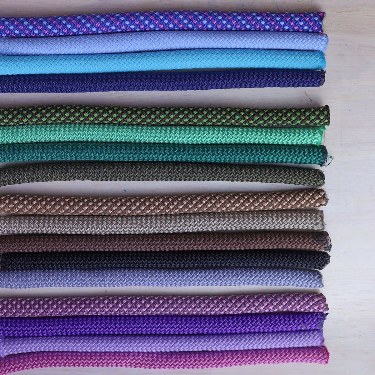 A collection of braided rope cords in various colors, including purple, blue, green, brown, black, and pink, arranged horizontally on a light wood background.